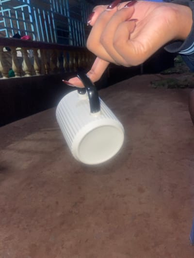 The image, set in Kisii Central ward, Kenya, depicts a close-up of a human hand holding a mug in a low-light outdoor or semi-outdoor environment. The hand, with dark skin tone, displays long, almond-shaped fingernails painted a dark reddish-brown with white tips. It holds a white ceramic mug with vertical ridges by its black handle, using a single finger (index finger) looped through the handle. The mug is oriented horizontally, its open end facing right. The background on the left features a light-colored balustrade or railing structure, behind which a building with blue walls or window frames is partially visible, with dark objects resting on the railing. The ground surface in the foreground and mid-ground is reddish-brown, appearing as compacted earth or an unpaved area with some textural variations. Faint green vegetation and disturbed ground are visible in the far background on the right. The overall illumination is dim, suggesting dusk or nighttime conditions.