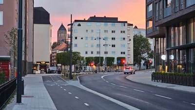 The image depicts an urban street scene at dusk in Linköping, Sweden. A silver car is driving on a curved asphalt road with white lane markings, including a bicycle lane indicated by bicycle symbols. On the right side of the road, a modern building with dark brick facade and large glass windows stands, featuring a black metal fence in front of it. On the left, a lighter colored building with multiple windows is visible, and further down the street, a tall tower with a distinctive roof is partially obscured by other buildings. Several streetlights are present, and traffic lights are illuminated red. The sky displays a soft gradient of pink and orange, indicative of sunset. There are no people clearly visible in the frame.
