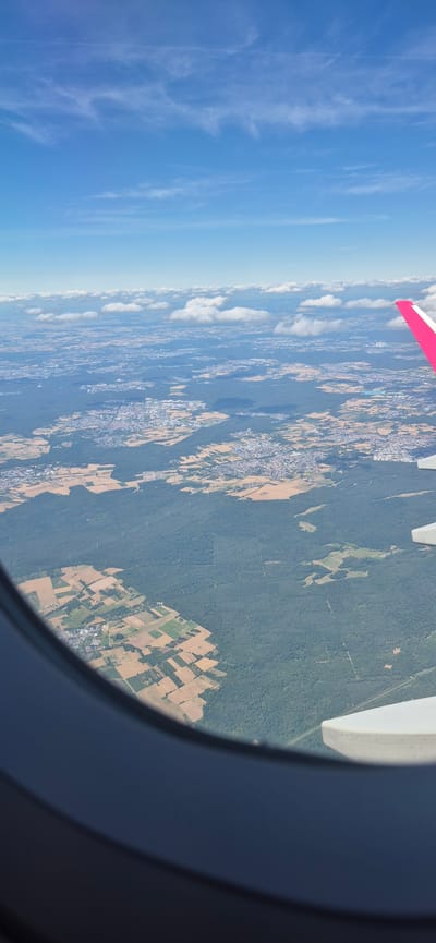 An aerial view from an airplane window shows a landscape dotted with agricultural fields, forests, and urban areas. The sky above is a bright blue with scattered white clouds. The airplane wing, with its pink tip, is visible on the right side of the frame. The ground below consists of a patchwork of green forests and lighter colored agricultural fields, some appearing to be recently harvested. Small towns and clusters of buildings are interspersed within the landscape. The perspective is from a high altitude, looking down at the earth. The location is identified as Spata-Loutsa Municipal Unit, Greece.