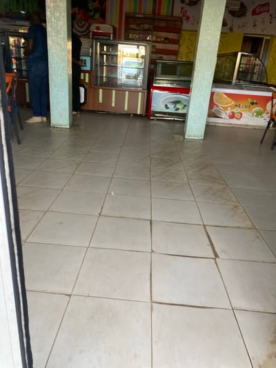 The image depicts the interior of an establishment, possibly a cafe or restaurant, with tiled flooring. In the background, there are display cases containing food items, likely pastries or ice cream, and refrigeration units. One refrigeration unit prominently features a "NEXUS" logo with an illustration of fruits and ice cream. A person is visible in the left of the frame, wearing a blue shirt and dark pants, standing near a counter. Another person is partially visible behind a pillar. The overall environment appears to be a commercial space in Barakin Lerawi, Nigeria, with decorative elements on the walls and shelves displaying products. The lighting suggests an indoor setting.