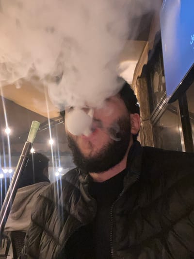 A person wearing a dark jacket exhales a large cloud of smoke, presumably from a hookah. The person has dark hair and fair skin. A hookah hose with a green tip is visible. The setting appears to be indoors with several lights. A screen or television is partially visible in the upper left corner. The image is rotated.