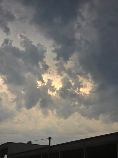 The image captures a dramatic sky with heavy, dark clouds partially obscuring the sun, which creates a luminous glow. The sunlight breaks through in patches, illuminating the clouds from behind and casting a warm, golden light. On the right side of the frame, a section of a dark, modern building structure is visible, with angled lines suggesting an overhang or a roof. A thin cable stretches from a pole attached to the building, disappearing into the sky. The overall scene suggests an impending storm or the aftermath of one, with the light breaking through the dense cloud cover. The time of day appears to be either early morning or late afternoon, given the low angle of the sun and the diffused light. The location is identified as Sofiivska Borshchahivka, Ukraine. There is no visible text in the image.