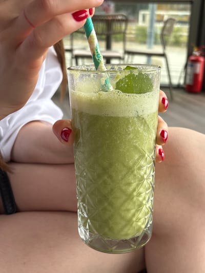The image shows a person holding a tall glass filled with a frothy green beverage, likely a smoothie or juice. The glass has a decorative, diamond-patterned texture. A striped straw, green and white, is inserted into the drink. The person's hands are visible, with bright red nail polish on their fingernails. One hand is holding the glass, and the other is near the straw, suggesting they are about to take a sip or are holding it in place.
The background is out of focus but provides some context. Through a window or open area, there are indications of an outdoor setting with greenery and possibly some structures. A red object, possibly a fire extinguisher, is visible in the upper left background. The lighting suggests it might be daytime, possibly with bright, natural light. The overall impression is one of refreshment and relaxation, perhaps during a warm day. There is no visible text in the image. The location context is Kragujevac, Serbia, but this information is not visually discernible from the image itself.