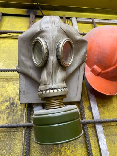 A close-up, eye-level shot shows a worn, light gray gas mask hanging against a yellow, textured surface. The gas mask has two round, glass eyepieces with metallic rims and a green, cylindrical filter attached to the bottom. To the right of the gas mask, partially visible, is an orange hard hat. The objects appear to be resting against or hanging on a structure made of rebar and metal bars. The background is a brightly lit, rough, yellow surface, possibly a wall or a large piece of equipment. The lighting is direct, casting faint shadows. The image does not provide any information about the city of Bishkek, Kyrgyzstan, or any specific location within it. There are no people or discernible actions in the scene.