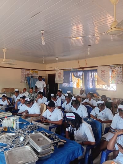 An indoor educational setting, likely a classroom or laboratory, is observed in Ejule, Nigeria. Approximately 25-30 individuals, predominantly young females, are present, dressed in white nursing uniforms with white caps featuring a red stripe. They are seated at blue tables draped with blue tablecloths, occupying blue plastic chairs. A male individual in a white t-shirt stands near the back, observing the group. The foreground tables display a large quantity of metallic medical instruments, including various forceps, clamps, scissors, retractors, kidney dishes, and stainless steel trays. A clear bottle containing a yellow liquid and a roll of white tape are also on a table. Many students are looking downwards, some engaging with mobile phones, while others appear to observe the instruments or notes. The room features light-colored walls with a horizontal dark stripe. Anatomical charts depicting human organs such as the liver, digestive system, and eye/ear are affixed to the back wall. Windows with metal grilles and blue curtains are visible. The ceiling is constructed from white PVC panels and is equipped with multiple exposed incandescent light bulbs and several white ceiling fans.
