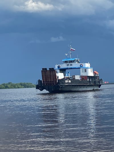 A barge is seen from the side, partially submerged in calm water. The barge is dark gray and has a white superstructure with blue accents. The text "СП-26" is visible in white on both the superstructure and the side of the barge. A small Russian flag is visible at the top of the superstructure. The barge appears to be carrying a load of what looks like industrial equipment, possibly scrap metal, on its deck. The sky is blue with scattered white clouds. The scene is likely set in the morning or early afternoon, given the lighting. The water is dark blue and reflects the sky and the barge. A thin line of green trees is visible along the horizon in the distance. The overall mood of the image is calm and serene.