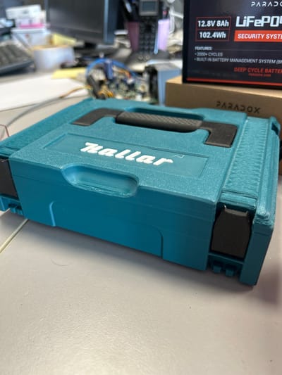 A teal-colored toolbox with a white "Makita" logo is placed on a light gray surface. To the right of the toolbox, a cardboard box with "PARADOX" printed on it is visible. Above the cardboard box, a black and red box with "PARADOX SECURITY SYSTEM" is positioned. The background is out of focus but appears to be an office environment with computer equipment and electronic components. The image was taken indoors, likely in a workshop or office.
