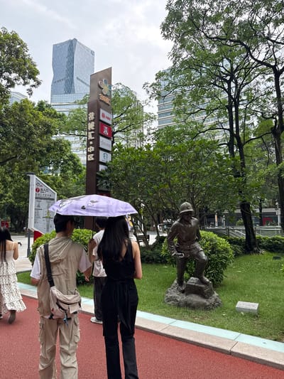 This image depicts a street scene in Tianhe District, China, on a cloudy day. Several people are present, some walking and others posing. A young woman in the foreground holds a purple umbrella adorned with cartoon characters. She is wearing a black sleeveless top and black pants. To her right, another individual is seen from the chest down, wearing a tan-colored outfit consisting of a short-sleeved shirt and loose pants, with a light-colored fanny pack worn across their chest. Further down, another person is partially visible, wearing a white dress with black polka dots.

In the background, a bronze statue of a worker is visible amidst lush green grass and bushes. Beyond the greenery, modern glass-fronted buildings rise into the sky, suggesting an urban environment. A prominent signpost displays various logos and Chinese characters, including "KFC" and what appears to be a map or directory with an "Entrance/Exit" marker that reads "Gate 7" and "Huachenghui Central District Atrium" in Chinese. The overall impression is of a public space within a bustling city, possibly a park or a pedestrian area adjacent to commercial buildings, with overcast weather contributing to a diffused light.