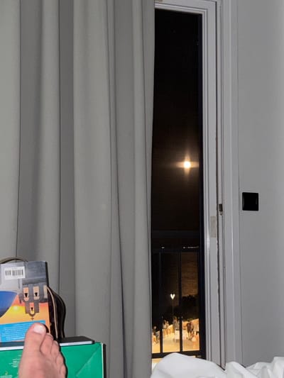 A partially drawn grey curtain obscures the left side of the image, revealing a white framed window on the right. Through the window, a dark night sky is visible with a bright, full moon positioned slightly off-center. Below the moon, a balcony railing and an illuminated outdoor area with several silhouetted figures can be seen. In the foreground on the left, a foot is visible, and to its right, a book with an illustration of buildings is propped up by a bag. The scene suggests an interior view from a room in Kassandra Municipal Unit, Greece, looking out at a moonlit night.
