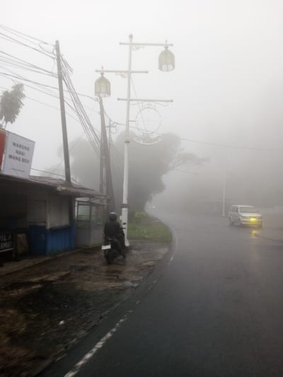 The image displays a heavily fog-obscured street scene. The scene is located in the city of Neerveli, Sri Lanka. A wet, two-lane asphalt road curves from the foreground right into the thick fog, with its left edge marked by a solid white line.
On the left side of the road, a single-story structure with a corrugated metal roof, a light-colored upper facade, and a blue lower section is visible. An overhead sign, white text on a red background, reads "WARUNG NASI WANG BEO". A person wearing a dark helmet and dark jacket is seated on a black motorcycle, parked on the damp concrete area in front of this building. Behind the building and motorcycle, multiple wooden utility poles are visible, supporting a dense network of overhead wires.
Centrally positioned near the road's curve is a decorative white streetlamp pole featuring two ornate lanterns and a circular sign below them that reads "AL BAASITH". The language on the signs, "WARUNG NASI WANG BEO" and "AL BAASITH", is Indonesian.
Further down the road on the right, partially visible through the dense fog, is a light-colored van with its headlights illuminated, appearing to travel towards the viewer. Trees and other distant objects are largely obscured by the pervasive fog.