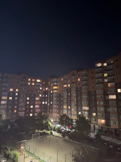 This media file captures a nighttime view of a large, multi-story residential building in Vinnytsia, Ukraine. The building is illuminated by numerous lit windows, suggesting that many apartments are occupied. The dark sky indicates it is nighttime, and there are no visible weather cues.
The scene depicts a typical urban residential setting. In front of the building, there is an outdoor recreational area that appears to include a small soccer goal, playground equipment, and some trees. The ground is paved. The perspective is from a slightly elevated position, looking down and across the building and the recreational area.
There are no clearly identifiable people or specific objects of interest apart from the building itself and the amenities in front of it. The activity taking place is simply the observation of the residential building at night. There are no strong emotional cues conveyed, beyond a general sense of an urban environment. No visible text is discernible in the image.