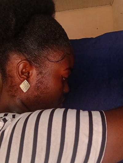 A close-up captures a person's head and upper body. The individual lies on a blue surface, possibly a pillow or bed, with their head turned sideways. A diamond-shaped earring adorns their ear. Their dark hair is styled in a large bun at the back. The person wears a striped shirt in black and white. The focus is on the person's facial profile and the details of their hair and earring. A wall is visible in the background.