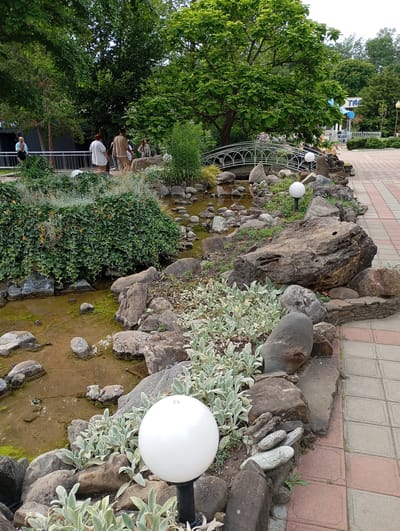 The image shows a well-maintained park area in Goryachiy Klyuch, Russia. A man and two women are walking along a paved path in the background, with several people visible further in the distance near a building with a sign that appears to say "TA". In the foreground, a rocky stream bed is adorned with lush green ivy and other low-lying plants. White spherical lamps are strategically placed amongst the rocks, illuminating the area. A decorative arched bridge spans the stream in the middle ground. The overall scene depicts a peaceful and scenic outdoor environment, likely a popular spot for recreation and relaxation.