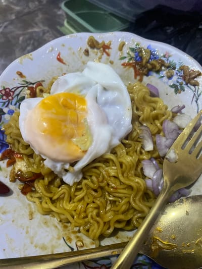 A close-up, overhead shot captures a plate of instant noodles topped with a fried egg and scattered red onion slices. The noodles are golden yellow and curly, mixed with chili and some unidentifiable ingredients. The fried egg, positioned slightly off-center, features a runny yolk. A fork and spoon, both appearing to be gold-colored, rest on the plate. The plate itself has a floral pattern with faded colors and is smudged with sauce and food residue, indicating it has been used. The background is blurred, but suggests a dark, possibly indoor setting with glimpses of green plastic containers. Based on the style of the plate, the presentation of the noodles, and the common availability of instant noodles with fried eggs and onions as a popular dish, the scene is likely located in a food stall or a casual eatery in North Jakarta, Indonesia.