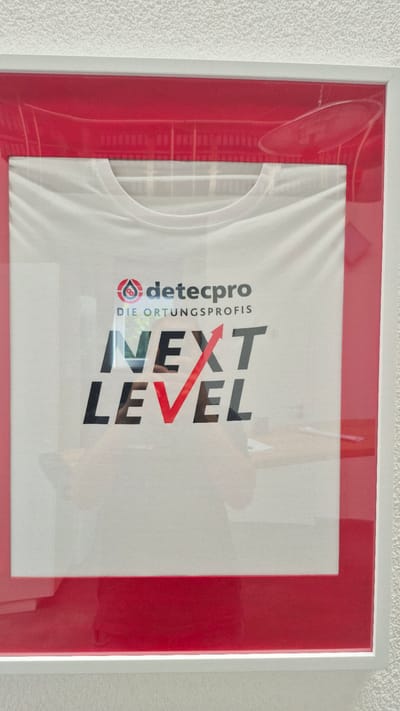 This image shows a white t-shirt framed with a red matting and a white frame. The t-shirt has a logo and text printed on it. The logo is a red circle with a stylized person inside, next to the text "detecpro". Below that, it says "DIE ORTUNGSPROFIS". Further down, the words "NEXT LEVEL" are printed in large black letters, with a red arrow forming the crossbar of the "T" in "NEXT" and extending over the "V" in "LEVEL". The t-shirt is displayed on what appears to be a wall in an indoor setting. The framing and display suggest it is a decorative item. There is a reflection on the glass of the frame, showing a person and possibly a room interior. This item is located in Muggensturm, Germany.