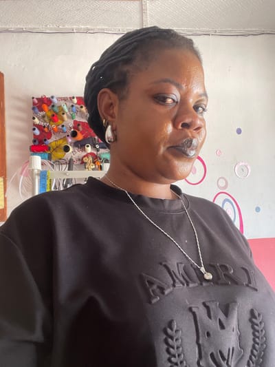 The media file is a portrait of a woman with dark skin and dark hair styled in braids. She is wearing a black sweatshirt with a textured design on the chest, featuring what appears to be lettering and ornate embellishments. A thin silver chain necklace is visible, with a small charm attached to the sweatshirt. She has large hoop earrings in her ears and is wearing dark lipstick.
The background suggests an indoor setting, possibly a crafting or sewing area. To the left of the woman, a wall is visible with a textured, patterned surface and some decorative circular elements in pink and blue. To her right, a display board is filled with spools of colorful thread, arranged in a seemingly organized manner. A blue measuring tape, showing markings "31 32", is also present. The lighting suggests it is daytime. The woman's expression is direct, with her head tilted, looking towards the camera.
The location context provided is Badagry, Nigeria, which adds a cultural and geographical dimension to the scene. While no specific time of day or weather conditions are explicitly depicted, the indoor setting and lighting imply a typical day. The overall impression is one of a person in their personal space, possibly engaged in or near a creative activity.