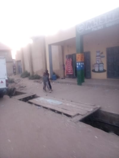 An outdoor scene in Rangaza, Nigeria, depicts a commercial street or pathway under bright, possibly overcast, daylight conditions. In the foreground, an open, dark trench or drainage ditch extends across the width of the image. Portions of this ditch are covered by makeshift wooden plank bridges or walkways. To the far left, the front portion of a white vehicle, possibly a van or small truck, is visible, featuring a red triangular reflector.
In the midground, two individuals are walking from left to right on the unpaved, dusty ground. One individual, appearing smaller, wears light-colored trousers and a dark top. The other, taller, wears dark clothing. Their specific features are indistinct due to motion blur or image quality.
The background features a row of light-colored, single-story commercial buildings. A prominent structure on the right has a dark green sign overhead displaying "M.ISH..." and "PAINT DEPOT" in light lettering. Below this sign, a green vertical pillar decorated with yellow, orange, and blue geometric patterns stands. To the right of the pillar, a mural on the building wall depicts a stack of paint cans. To the left of the pillar, an open doorway or display area reveals a brightly patterned garment, predominantly pink/red with black designs, hanging for display. Further left along the buildings, additional doorways and sections of plain, light-colored walls are visible. Sparse vegetation grows intermittently along the base of the buildings. The ground throughout the scene appears unpaved and dusty.