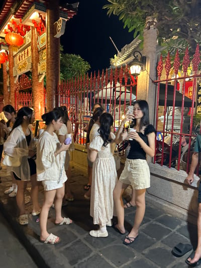 In Hội An Ward, Vietnam, a group of young women are gathered at night in front of a vibrantly decorated building, possibly a temple or a traditional establishment, characterized by red ornate fences and glowing lanterns. The architectural style suggests a strong East Asian influence. The women, dressed in light-colored casual summer clothing, are interacting with each other. One woman in the foreground is holding a cup and a phone, appearing to take a selfie. Another woman next to her is holding a purple cup. The ground is paved with stone tiles. The scene is illuminated by artificial lights, creating a warm and lively atmosphere.