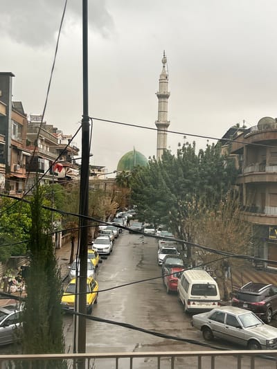 The image displays an urban street scene in Sarouja Municipality, Syria, under an overcast sky, with the asphalt street surface appearing wet. A prominent mosque featuring a green dome and a tall, slender, light-colored minaret is visible in the background. Multi-story buildings, likely residential or mixed-use, with balconies, line both sides of the street, which slopes downwards from background to foreground. Numerous vehicles, including various sedans (one yellow, one light grey in the foreground), a white van, and an SUV, are parked along both curbs of the street. A large black utility pole with multiple black overhead wires extends across the central and left portions of the foreground, partially obscuring the view. Trees with green foliage and some with bare branches are present along the street, notably a tall, slender evergreen tree on the left. At least one pedestrian, dressed in dark clothing, is visible walking down the street in the mid-ground. A collection of debris or refuse is visible on the left side of the street, partially hidden by vegetation. A metal railing is present in the immediate foreground, indicating the viewpoint is from a balcony or elevated position.