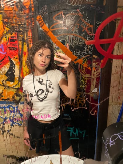 A young woman with curly brown hair takes a selfie in a mirror covered in graffiti. She is wearing a white t-shirt with a black beaded necklace and black shorts. Her left arm is tattooed. The background is a wall covered in various graffiti tags and drawings, including what appears to be an anarchy symbol. The environment is cluttered and appears to be in an urban setting.