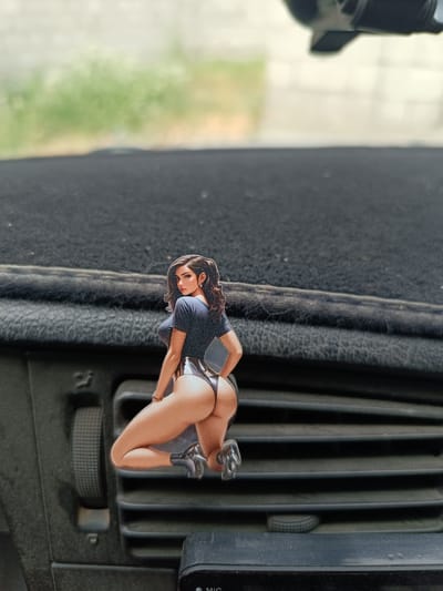 An illustration of a woman in a dark top and skimpy bottoms is attached to a car's air vent. The woman is depicted from behind, with her head turned to face the viewer. She has long, dark hair and is wearing earrings. Her pose is suggestive. The air vent is black and has horizontal slats. To the left of the vent is a circular dial with ridges, and to the right of the vent is a small black device with the word "MIC" visible. The background of the image is blurred, showing a patch of green foliage and a light-colored surface. The overall scene appears to be inside a vehicle.