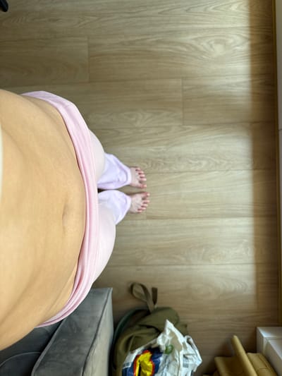 A top-down, close-up view focuses on a person's midsection and legs, clad in pale pink leggings, standing on a light-colored wood-laminate floor. The person's bare feet are visible, positioned slightly apart. The environment appears to be indoors, possibly a residential setting, with a soft gray sofa partially visible in the lower left corner and a pile of bags and other items in the lower right. The lighting suggests natural light entering from the right side of the frame, casting a shadow on the floor. There is no indication of the city of Krakow, Poland, or any specific urban context in the image.