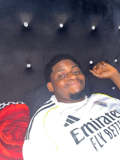 This image depicts an adult male indoors in Ube, Nigeria. The male, with dark skin, short, dark, curly hair, and a goatee-style beard, is positioned against a dark, possibly velvet, tufted headboard or wall panel adorned with clear, faceted buttons arranged in a grid pattern. He is wearing a white short-sleeved sports jersey featuring black Adidas stripes on the shoulders and a Real Madrid crest on the left chest. The jersey prominently displays the sponsor text "Emirates FLY BETTER" in black lettering. His left hand is partially visible in the upper right quadrant of the frame, with fingers resting against the dark background. A portion of a patterned pillow or blanket, displaying a large red rose design on a red background, and a small section of black and white polka dots, is visible in the lower-left corner of the frame. The male exhibits a slight smile and is looking directly towards the camera.
