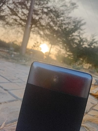 A dark, rectangular object with rounded edges dominates the right side of the frame, possibly a book or tablet case. It features a shiny, reddish-brown upper section contrasting with a matte black surface below. The background is blurred, revealing trees and a bright, diffused light source, likely the sun. The scene appears to be outdoors, possibly during sunrise or sunset, and a textured surface is partially visible beneath the object.