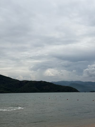 The image shows a cloudy day over a body of water in Hoa Thang, Vietnam. The water is calm with some ripples and a patch of white foam on the left side, possibly from a boat or wave. Numerous orange buoys are scattered across the water's surface. In the background, a dark green, densely vegetated hill slopes down to meet the water. Further in the distance, hazy, layered mountains recede into the horizon under a gray, overcast sky. The shoreline, visible in the lower right, is a sandy beach. No people are present in the scene.