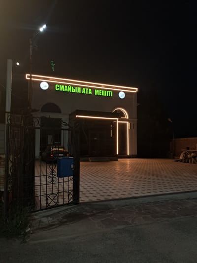 This is a night-time photograph taken outdoors, possibly in Shymkent, Kazakhstan. The main subject is a building with illuminated signage. The building appears to be a mosque, as indicated by the Arabic script on the sign and a crescent moon motif. The sign, written in green neon letters, reads "СМАЙЫЛ АТА МЕШІТІ" which translates to "Smail Ata Mosque".

The building has a modern architectural style with large windows and glowing orange neon accents along its facade, outlining geometric shapes and architectural features. In front of the building, there is a decorative metal fence and a small blue mailbox. A portion of a vehicle is visible behind the fence.

The ground in front of the building is paved with a patterned tile, and to the right of the paved area is a rougher, textured surface that appears to be a road or sidewalk. The overall atmosphere is dark due to the time of day, with artificial lighting from the building and a street lamp creating a stark contrast. The weather is not discernible from the image. The scene appears quiet and still, suggesting a typical evening outside a place of worship.