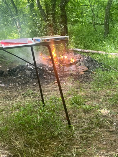 The image shows a fire burning outdoors, surrounded by charred earth and green vegetation. A metal stand is positioned near the fire. The environment suggests a natural setting, with trees visible in the background.