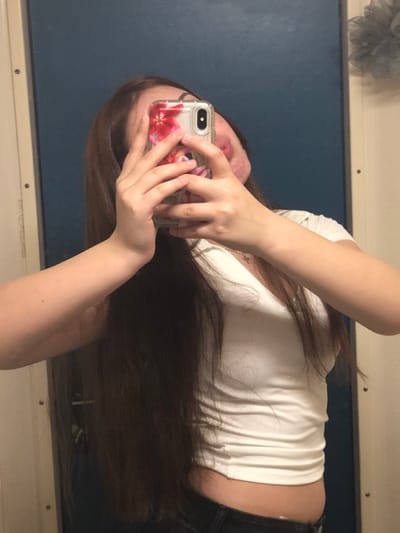 The image displays a mirror selfie of a person, likely female, with long, dark brown hair. The image is oriented on its side, with the person's head to the left and feet to the right if viewed upright.

The person is holding a smartphone with both hands, obscuring most of their face. A clear phone case with a vibrant floral design, featuring prominent red or pink flowers, is visible on the phone. Part of the person's mouth and chin are visible beneath the phone, showing pink-colored lips. A small, delicate silver-colored necklace with a tiny "X" or cross-shaped pendant is worn around the neck.

The person is wearing a form-fitting, white ribbed crop top that reveals a portion of their midriff, and dark-colored bottoms, possibly jeans or pants. Their long, straight, dark brown hair cascades down their back and across their arm.

The background is a reflection of a room in a mirror. A dark blue or gray surface, possibly a wall, occupies most of the background, framed by lighter, off-white or cream-colored borders at the top and bottom, which could be part of a wall or furniture. In the top left corner of the reflected background, a blurry, fluffy gray object, possibly a bath puff or decorative item, is visible. The reflection suggests an indoor setting. No specific time of day or emotions are discernible. No text is explicitly visible in the image.