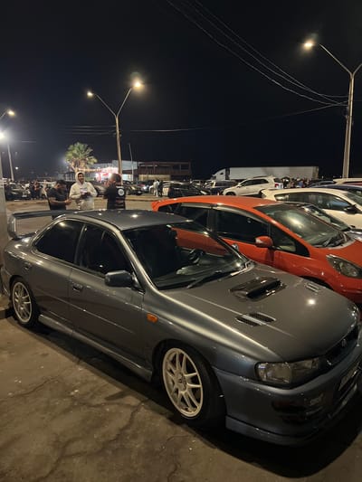 The image depicts an outdoor scene at night, likely a parking lot or gathering area, in the city of Arica, Chile. A grey Subaru Impreza WRX STI, a performance-oriented compact car, is prominently featured in the foreground, angled towards the viewer's left. The car has a large rear wing and white aftermarket wheels. To the right of the Subaru, an orange compact car, possibly a Honda Fit, is visible. Several other vehicles are parked behind these cars, including a white truck with a trailer, and various sedans and SUVs.

In the mid-ground, three men are standing and interacting. One man, wearing a white hoodie, is gesturing with his hands and appears to be speaking. Another man, in a black t-shirt with graphics, is looking towards the man in the hoodie. A third man, also in a black t-shirt, is standing to the left, holding a phone. The lighting in the scene comes from streetlights, creating distinct pools of illumination and casting shadows. In the background, there are glimpses of buildings, a palm tree, and possibly food stalls or other commercial structures, suggesting an urban or recreational environment. The ground appears to be asphalt or concrete, with visible cracks. The overall atmosphere suggests a casual social gathering or car meet-up.