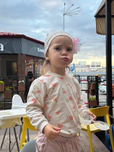 The media file shows a young child, likely a toddler, sitting at an outdoor cafe. The child is wearing a cream-colored long-sleeved shirt adorned with pink hearts and green stems, a light-colored beanie with a pink and white flower, and pink pants. The child holds a small plastic container with a lid, possibly for food, in their right hand. The background features a blurry urban scene with buildings and vehicles, suggesting a daytime setting. There is a yellow chair visible, indicating a seating area at the cafe. Partially visible text on a building in the background reads "КАФЕ" (Cafe in Russian). The lighting suggests it might be late afternoon or early evening, with overcast skies. The child's facial expression is somewhat ambiguous, with a slight pout. The overall scene depicts a casual moment at a cafe, with the focus on the child.