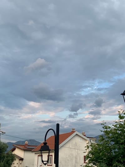 The image captures a cloudy sky over a residential area in Radovići, Montenegro. In the foreground, parts of houses with red tiled roofs are visible, along with a black lamppost. A lush green tree branches into the frame from the right. In the background, mountains are silhouetted against the sky, and a small airplane is visible in the upper left quadrant of the image. The clouds are a mix of grey and white, with hints of blue sky peeking through in some areas. The overall impression is one of a slightly overcast day in a coastal Montenegrin town.