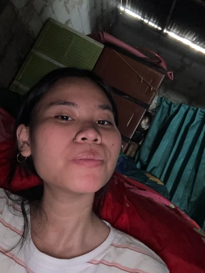 A selfie taken from a low angle shows a young woman lying down with her head propped up. She has dark hair and is wearing a white t-shirt with pink stripes and a hoop earring in her left ear. Her expression is neutral, with her mouth slightly open.
The background appears to be an indoor space with a rough ceiling and walls, possibly a storage area or makeshift dwelling. In the upper left, a green rectangular object is visible, possibly a mattress or a folded piece of fabric. To the right of that, a dark reddish-brown piece of furniture with latches is present. Behind the woman, a dark teal curtain is drawn, with some fabric peeking out from behind it. Sunlight streams in from gaps in the ceiling, creating bright lines. The overall impression is of a dimly lit, somewhat cluttered space.
The image's metadata suggests it was taken in East Jakarta, Indonesia, on May 19, 2024, at 10:19 AM.