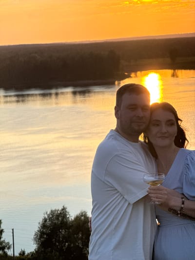 A couple embraces during sunset by the water. The woman, with dark hair, leans her head on the man's shoulder. She holds a glass of white wine in her right hand. She wears a light blue top and a bracelet on her left wrist with several charms. The man, with short brown hair, wears a white t-shirt and has his arm around her. They appear to be in a romantic and peaceful moment. The background shows a body of water reflecting the warm orange and yellow hues of the setting sun. Trees are visible on the right side of the frame, silhouetted against the sky. The overall mood is serene and intimate, capturing a beautiful evening in Ольховка, Russia.