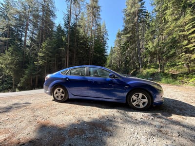 A blue four-door sedan is parked on the side of a road in a forest near Bansko, Bulgaria. The car is facing to the right of the frame, with its left side visible. The road is paved and curves to the left. The ground next to the road is covered in gravel and pine needles. Tall pine trees line both sides of the road, with a clear blue sky above. The sun is shining, casting shadows on the ground. There are no people visible in the image.