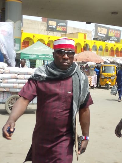 The image depicts a daytime street or market scene in Fagge C, Nigeria. In the immediate foreground, an adult male, appearing middle-aged, is walking forward, facing the camera. He wears a red and white striped traditional cap, dark sunglasses, a grey and black patterned scarf, and a short-sleeved maroon tunic with a breast pocket. His right hand holds a small dark blue object. His left hand holds a flat, light brown rectangular object and a dark wallet/bag, and he wears a red, white, and blue striped wristband.
In the midground to the left, a wooden two-wheeled cart is laden with numerous white sacks. Behind this, a green tent with partial white lettering, possibly reading "May your table always," is visible. A person is partially obscured behind the tent.
In the midground to the right, a yellow three-wheeled vehicle (auto-rickshaw) is parked. An orange parasol provides shade over an indistinct stall. Another individual in a blue garment is seen walking away, carrying a black bag.
The background features a long, yellow building with multiple arched entrances and red-orange doors, suggesting a market or commercial structure. Above this building, two large billboards display "PASSION ENERGY DRINK" with the slogan "MAKE LIFE SWEET WITHOUT PASSION." The ground is an unpaved or dusty surface.