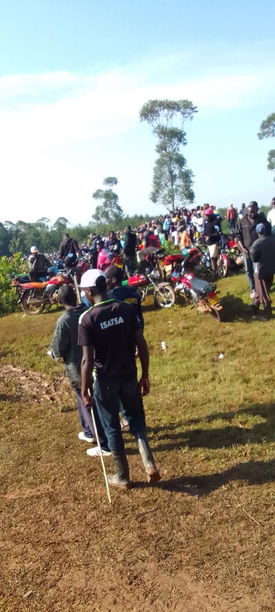 The scene is a crowded gathering of people near a dirt road in Isukha ICHINA, Kenya. A large group of people are standing along the dirt road and on the grassy slope. A number of motorbikes are parked in a line along the road. Two men wearing black, blue, and white clothes are standing in the foreground. The man on the right is wearing a black shirt with white lettering that reads "ISATSA". Both men are looking toward the group of people. The ground is covered in brown grass and dirt. The sky is clear with a few white clouds. There are trees in the background. The area is sparsely populated. The scene is peaceful and orderly.