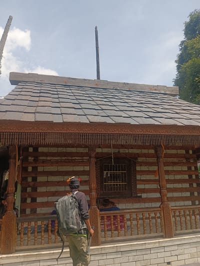 The image depicts the exterior of a traditional wooden and stone building with a slate tile roof. The building features ornate wooden carvings along the eaves and posts. A man wearing a backpack and a traditional hat stands with his back to the camera, facing the building. He is holding a long object, possibly a walking stick or trekking pole, in his right hand. The building has a porch area with a wooden railing and a barred window. The surrounding environment includes trees and a cloudy sky. The provided crops highlight details of the roof, the wooden carvings, and the man's attire and backpack. The scene is not identifiable as being in Gojra, India.