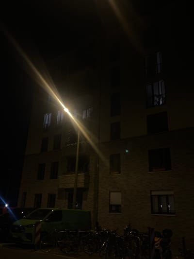 The image depicts a building in Berlin, Germany, at night. A bright street lamp in the foreground casts a strong glare across the scene, illuminating a portion of the brick building. Several windows are visible on the building's facade, some appearing dark and others with faint interior light. Below the building, in the foreground, a green van and several bicycles are parked. The environment appears to be a residential or commercial street.