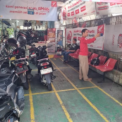 The scene is a motorcycle shop in Bogor, Indonesia, located at latitude -6.562 and longitude 106.795. The shop is lined with Honda motorcycle parts advertisements. There are four motorcycles parked in front of the shop. Three of the motorcycles are black, and the other is silver. The black motorcycles have license plates: 2128 DZ, 7100 Z, and 5L27 AAW. The silver motorcycle has a license plate that is not visible. A man is standing in front of the shop and pointing at an ad for Honda parts. He is wearing a red shirt and brown pants. There are three red chairs in front of the shop. A man in a blue shirt and khaki pants is sitting on one of the chairs. The shop's entrance is lined with a yellow and green pattern. The floor is gray. The shop's entrance is open. There is signage above the entrance, stating "Cari Aman", and advertising Honda genuine parts.