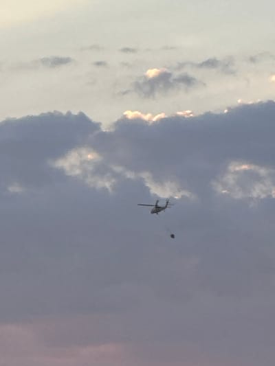 The media file depicts a clear sky with scattered clouds, suggesting daytime, possibly late afternoon given the warm glow on some clouds. The setting appears to be outdoors, with the vast sky dominating the frame.
The primary subject is a helicopter in mid-flight. It is a dark silhouette against the lighter sky, and its rotors are visible, indicating it is actively flying. To the right of the helicopter, a small, dark, spherical object is also visible in the air. Its nature is unclear from the image alone.
The activity taking place is aerial. The helicopter is in motion, and the presence of the separate spherical object might suggest an operation or a specific event, though this is speculative.
Notable details include the bright, sunlit edges of some clouds, contrasting with the darker, more shadowed parts. The sky itself transitions from a bright, almost white hue on the left to a softer blue-grey on the right.
There is no visible text in the image. The location context provided is Fethiye, Turkey, which, combined with the sky and clouds, offers a potential geographical setting. The overall mood conveyed is one of open space and aerial activity.