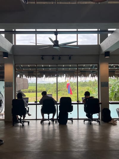 The image depicts the interior of a co-working space with a large window offering a view of a lush, tropical landscape. Three individuals are seated at a long desk, each working on a laptop. The person on the left is wearing a light-colored shirt and has their feet bare. The person in the middle is wearing a patterned shirt, also with bare feet. The person on the right is wearing a dark tank top. A Filipino flag is visible flying in the distance. The co-working space features a large ceiling fan and minimalist furniture. Signs indicating "QUIET ZONE" are posted on the walls. The overall ambiance suggests a relaxed yet productive environment. The co-working space is located in General Luna, Philippines, a popular tourist destination known for its surfing beaches and vibrant atmosphere.