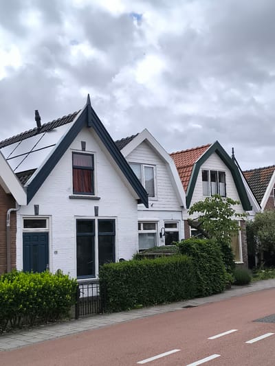 A photograph captures a row of charming gabled houses lining a residential street under an overcast sky in what appears to be Alkmaar, Netherlands. The weather is cloudy, with large, light grey clouds dominating the upper portion of the frame, suggesting a dull or cool day. No people or active events are visible.
In the foreground, a reddish-brown road or path with two sets of white dashed lines runs parallel to the houses, likely indicating a dedicated bike lane. A grey paved sidewalk separates the path from neatly trimmed green hedges that front the properties.
The houses display a mix of architectural styles, predominantly featuring prominent gables. The most prominent house on the left has a white brick facade with dark blue trim along its gabled roof. Solar panels are installed on the left slope of its roof. A dark blue multi-paneled door is visible, next to which is the house number "98". Below the main gable window, which has red curtains, are two large dark-framed windows on the ground floor, one of which is partially obscured by a black metal gate and the hedge.
To its right, the next house has a white clapboard siding on its gable and a contrasting red-tiled roof. A window with white blinds is centered in its gable. On the ground floor, the house number "96" is visible next to a white door, above which a black wall lamp is mounted. Further to the right, another similar house features white clapboard siding with dark green gable trim and a red-tiled roof, with a window visible in its gable. A small, leafy tree grows in front of this house, partially obscuring the lower part of its facade. The houses continue to the right, with partial views of brick and dark-tiled roofs.
