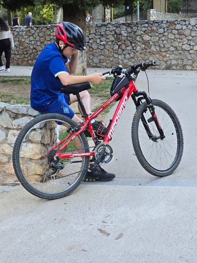 A young person wearing a blue shirt, black shorts, a red and black helmet, and white earbuds is sitting on a stone wall next to a red bicycle. The person is looking down at their hands, which are holding a mobile phone. The bicycle is a mountain bike with knobby tires and a black water bottle cage. In the background, there is a stone wall, trees, and other people walking. The scene appears to be in a park. The location in the city of Cornellà De Llobregat, Spain, is a public park with a paved walkway and a natural stone wall.