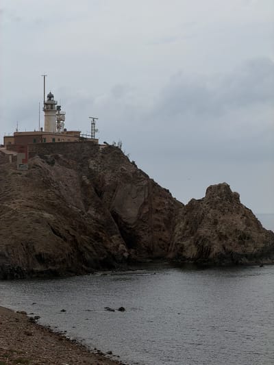 This is a landscape photograph taken in Níjar, Spain. The image depicts a rocky cliff face overlooking the sea under an overcast sky. The cliff is steep and weathered, with layers of rock visible. On the cliff, there is a lighthouse with a white cylindrical tower and a smaller building attached. Several communication antennas and dishes are mounted on towers near the lighthouse. The sea is calm and grey, with the texture of ripples visible on its surface. The shoreline is a mix of rocks and sparse vegetation. The lighting suggests it is daytime, likely overcast, as there are no strong shadows. The scene is quiet and evokes a sense of solitude and rugged natural beauty. No people are visible in the frame. No visible text is present in the image.