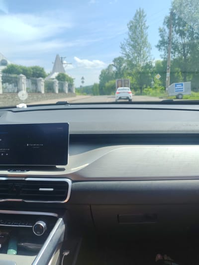 The image shows a view from inside a car looking forward. The dashboard of the car is visible, including a touchscreen display showing music playback. The car is on a road with a white fence and buildings on the left, and trees and a utility pole on the right. Further down the road, a white car is driving away from the camera, and a small trailer is parked on the right side of the road. The sky is blue with some clouds, and the overall lighting suggests it is daytime. The image appears to have been taken in the city of Mezhdurechensk, Russia.