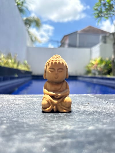 A small, light brown wooden Buddha statue is the central subject, positioned horizontally on a textured gray surface. Behind the statue, a blue structure is visible, possibly a part of a pool or similar feature. In the background, blurred elements suggest foliage, a building with a gray roof, and a blue sky with clouds. The location is likely Dalung, Indonesia, based on the user's prompt. No people, interactions, or specific actions are discernible in the image.